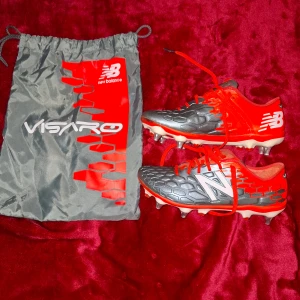 New Balance Visaro fotbollsskor röd/grå - Säljer ett par New Balance Visaro fotbollsskor i storlek 40.5. Skorna är grå med röda detaljer och har en cool hexagonstruktur på ovandelen. De har snörning och är tillverkade i syntetmaterial, perfekta för dig som vill sticka ut på planen. Medföljer originalpåse.