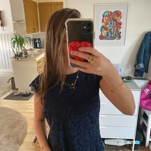 Mörkblå spetsblus med kort ärm - Säljer en mörkblå blus i spets med fina detaljer och korta ärmar. Blusen har en feminin och elegant look med vackert mönstrad spets och lätt transparenta partier vid axlarna. Perfekt att styla med jeans eller kjol för en snygg outfit.