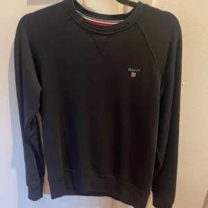 Svart sweatshirt från Gant - En stilren svart sweatshirt från Gant med diskret logga och broderad sköld på bröstet. Tröjan har rund halsringning, långa ärmar och ribbade muddar. Perfekt för en avslappnad och snygg look.