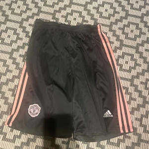  Manchester United shorts - Snygga svarta Manchester United shorts från Adidas med rosa ränder längs sidorna och klubbens emblem på ena benet. Perfekta för fotboll eller träning, med elastisk midja och sportig look. Materialet är mycket andningsbart 
