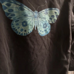 Svart hoodie - Svart hoodie från The Cove Company med stor blå fjäril på bröstet. Klassisk känguruficka, snörning i huvan och mjukt material. Perfekt för dig som gillar unika tryck och chill stil.