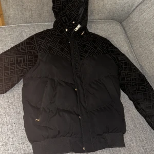 Svart pufferjacka Glorious Gangsta S - Svart pufferjacka från Glorious Gangsta i storlek S. Jackan har ett snyggt geometriskt mönster på axlar och huva, dragkedja framtill och broderad logga. Materialmix med vadderad polyester och detaljer i sammet. Perfekt för kalla dagar.