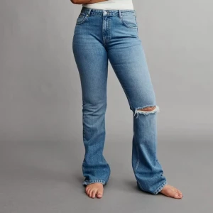 Blå bootcut jeans från Gina Tricot - Säljer ett par ljusblå bootcut jeans från Gina Tricot, modell Perfect Jeans. De har hög midja, en snygg sliten detalj med hål på ena knät. Jeansen är i mjukt denimtyg 