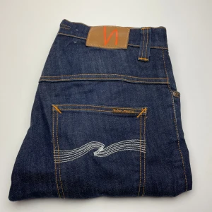 Mörkblå jeans från Nudie Jeans - Snygga mörkblå jeans från Nudie Jeans med klassiska orange sömmar och ikonisk broderad bakficka. Jeansen är i rå denim och har en rak passform som funkar till det mesta. Materialet är 100% bomull för en autentisk jeanskänsla.