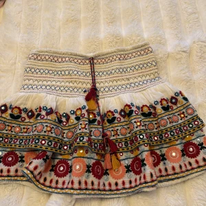 Zara kjol - Superfin kortkjol med broderade blommor och spegeldetaljer i färger som röd, gul, blå och rosa. Kjolen har en bred, smockad midja och snörning med tofsar framtill. Materialet känns som bomull och kjolen har volangkant för extra vibe.
