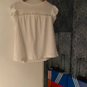 Vit blus med spets och volang - Säljer en söt vit blus med broderade detaljer och volang vid axlarna. Blusen har spetsband över bröstet och är tillverkad i lätt bomullstyg. Perfekt för dig som gillar romantisk och luftig stil. Passar till både jeans och kjol.