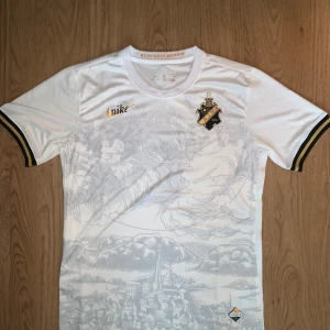 Vit AIK fotbollströja Nike S - AIK fotbollströja i vit Dri-Fit från Nike, med snyggt tryck av Stockholmsmotiv i grått över hela framsidan. Guldiga detaljer på logga och Nike-märke, samt svarta och guldiga ränder på ärmsluten. Rund hals och korta ärmar. Perfekt för dig som älskar AIK och stilrena matchtröjor.