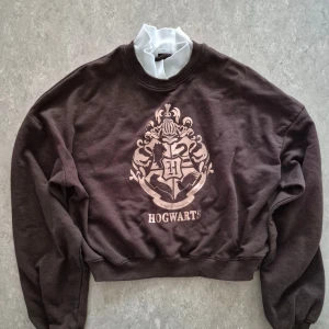 Tröja - Mysig brun sweatshirt från H&M med Hogwarts-tryck i silver på bröstet. Tröjan har rund hals, långa ärmar och ribbade muddar. Perfekt för dig som älskar Harry Potter och vill ha en chill och snygg look.