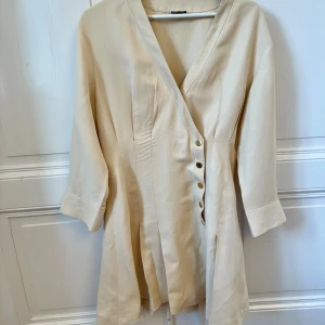 SANDRO Beige omlottklänning med knappar - Snygg beige kortklänning med omlott-design och markerad midja. Klänningen har v-ringning, trekvartsärm och dekorativa guldknappar längs framsidan. Tillverkad i ett mjukt tyg som ger en lyxig känsla och passar perfekt till vår och sommar.