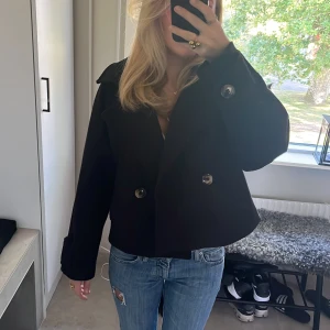 Svart kort kappa med knappar  - Säljer en svart kappa med bred krage och dubbelknäppning framtill. Jackan är croppad och har stora, bruna knappar både fram och vid ärmsluten. Perfekt för dig som vill ha en klassisk men trendig look till hösten elele vinten.💗