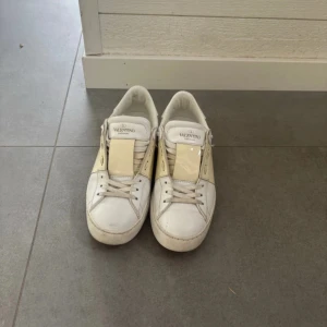 Valentino Garavani vita sneakers - Snygga vita Valentino Garavani sneakers med beige detalj över vristen och ikoniska nitar baktill. Skorna har snörning och är tillverkade i skinn med platt sula och rund tå. Perfekta för dig som vill ha en stilren och lyxig look.
