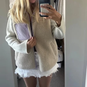 Beige fleeceväst med lila detaljer - Mysig och oversized beige fleeceväst med lila dragkedja och lila bröstficka. Västen har en mjuk teddylook och praktiska fickor på sidorna. Perfekt att slänga över en hoodie eller t-shirt för en chill och trendig stil.