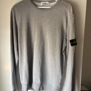 Grå sweatshirt från Stone Island - Snygg grå sweatshirt från Stone Island med klassisk rund hals och lång ärm. Tröjan har den ikoniska loggan på vänster ärm  Perfekt för en clean och stilren look. Alla lappar finns kvar för att scanna och bekräfta äktheten ✅ 