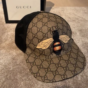 Gucci keps med bi och GG-mönster - Snygg keps från Gucci med klassiskt GG-mönster i beige och brunt, stor broderad bi framtill och svart mesh på sidorna och bak. Justerbar rem med grönt och rött band. Perfekt för dig som vill sticka ut med lyxig streetstyle.