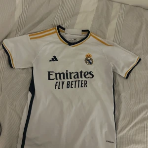 Real Madrid Adidas fotbollströja M - Säljer en officiell Real Madrid fotbollströja från Adidas i storlek M. Tröjan är vit med marinblå och gula detaljer, korta ärmar och klassisk v-ringning. Materialet är lätt och andas, perfekt för träning eller match. AEROREADY-teknologi för extra komfort.