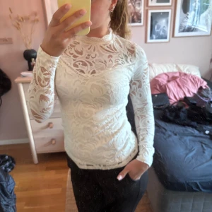 Vit spetsblus från Vila - Supersnygg vit spetsblus från Vila med långärmad design och hög hals. Blusen är genombruten med vackert spetsmönster och har en figurnära passform. Perfekt att styla med jeans eller kjol för en trendig look.