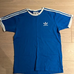 Blå Adidas t-shirt med vita ränder - Snygg blå t-shirt från Adidas med klassiska vita ränder längs axlarna och ärmarna. T-shirten har vit krage och ärmslut samt Adidas-logga på bröstet. Perfekt för en sportig look och chill vardagsstil.