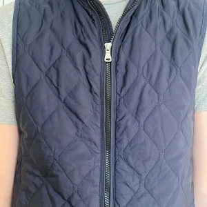 Mörkblå quiltad dunväst Oscar Jacobson - Snygg mörkblå quiltad dunväst från Oscar Jacobson med dragkedja framtill. Västen har ett stilrent rutmönster och är perfekt att slänga över en hoodie eller t-shirt. Materialet är mjukt och lätt, vilket gör den skön att bära under kyliga dagar.