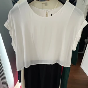 Vit och svart kortklänning med rund hals - Snygg kortklänning i vit och svart med rund hals och korta ärmar. Överdelen är vit och har ett lätt, luftigt tyg som ger en fräsch känsla. Kjoldelen är svart och kontrasterar snyggt mot toppen. Perfekt för dig som gillar stilren och enkel design.