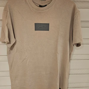 Beige t-shirt från Icono relaxed fit - Snygg mullvadsfärgad t-shirt från Icono i relaxed fit. T-shirten har ett svart rutigt tryck med logga på bröstet och klassisk rund hals. Tillverkad i mjuk bomull och har korta ärmar. Perfekt för en chill och stilren look.