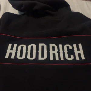 Svart HOODRICH hoodie med röda detaljer - Svart hoodie från HOODRICH med stor vit logga på ryggen och tunna röda linjer som detalj. Tröjan har huva med snörning och metallringar med HOODRICH-tryck. Perfekt för dig som gillar streetwear och vill sticka ut. köpte den för 1100kr från jd