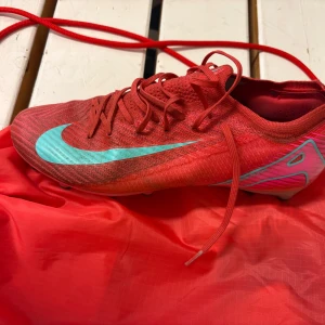 Nike fotbollskor - Snygga röda Nike fotbollsskor med turkosa swoosh och detaljer. Skorna har snörning och mönstrad ovandel i syntetmaterial. Sulan har dobbar i turkos och röd för bästa grepp på planen. Perfekta för dig som vill sticka ut på matchen!