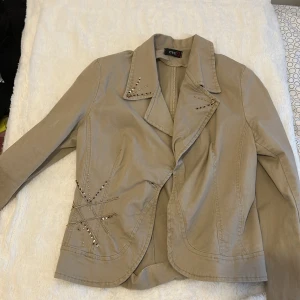 Beige kavajjacka med paljettdetaljer - Stilren beige kavajjacka från eve21 med snygga paljettdetaljer på kragen och vid ena höften. Jackan har klassisk krage, markerade axlar och är helfärgad. Perfekt för dig som vill sticka ut lite extra med detaljer.