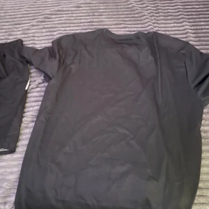 Svart Nike t-shirt med vit logga - Svart t-shirt från Nike med korta ärmar och vit swoosh-logga på bröstet. Sportig design med vita detaljer vid axlarna. Tillverkad i lätt polyester som andas, perfekt för träning eller chill. Klassisk rund hals och loose fit.