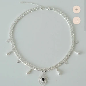 Silverhalsband med pärlor och hjärta från WOS - Stilrent halsband i silverfärgad metall med chunky kedja och hängande detaljer. Dekorerat med små pärlor och hjärtformade berlocker, samt en solinspirerad berlock med svart hjärta i mitten. Perfekt för att ge din look en cool edge.