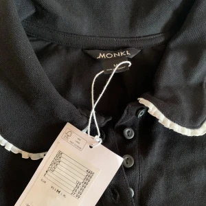 Svart blus med vit volangkrage Monki - Supersöt svart blus från Monki i bomull med korta ärmar och klassisk krage. Kragen har en vit volangkant som ger en gullig touch. Knappslå framtill med flera svarta knappar. Perfekt för dig som gillar en stilren men lekfull look.