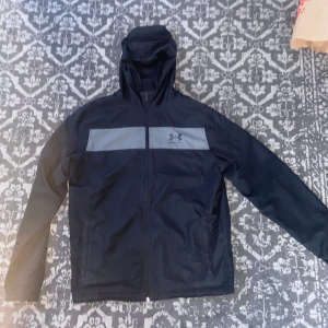 Svart vindjacka från Under Armour - Svart vindjacka från Under Armour med grått band över bröstet och logga. Jackan har huva, hel dragkedja och meshfoder på insidan. Perfekt för blåsiga dagar och sportiga outfits.