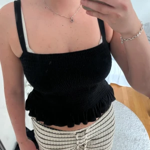 Svart smockad linnetopp med volang - Supersöt svart linnetopp med smockad struktur och breda axelband. Toppen har en volangkant nedtill som ger en gullig touch och sitter tajt men bekvämt. Perfekt att styla med jeans eller kjol för en trendig look.