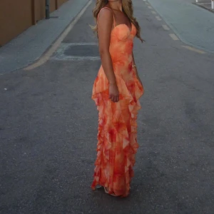 Lång orange volangklänning med öppen rygg - Säljer en långklänning i orange och ljusa toner med volanger och smala axelband. Klänningen har öppen rygg och ett blommigt mönster, perfekt för sommarkvällar. Materialet känns lätt och luftigt, och modellen är figurnära upptill med volanger nedtill.