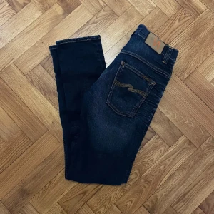 Mörkblå jeans från Nudie Jeans - Snygga mörkblå jeans från Nudie Jeans med klassisk femficksdesign och kontrastsömmar. Jeansen har raka ben och subtila slitningar framtill för en cool look. Tillverkade i kraftigt denimtyg med Nudie-logga på bakfickan.