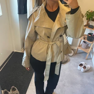 Kort beige trenchcoat med bälte - Säljer en snygg kort trenchcoat i beige med dubbelknäppning, bred krage och axelklaffar. Jackan har bälte i midjan och stora knappar framtill. 