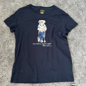 Mörkblå Polo Bear t-shirt Ralph Lauren - Mörkblå t-shirt från Ralph Lauren med Polo Bear-tryck framtill. Klassisk rund halsringning och korta ärmar. Tillverkad i mjuk bomull för en skön känsla. Perfekt för dig som gillar stilrena plagg med en twist.