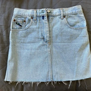 Ljusblå jeanskjol från Vans - Ljusblå jeanskjol från Vans med rå, fransig nederkant och klassiska femficksdetaljer. Kjolen har bälteshällor, knapp och dragkedja framtill samt en liten broderad sneaker på fickan. Perfekt för en chill och avslappnad stil. Storleken står inte i kjolen, men är ungefär i storlek XS.🩵