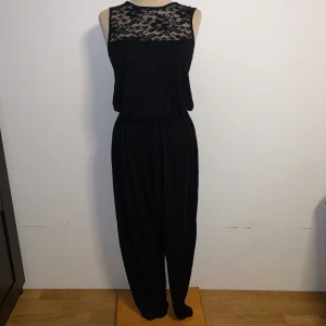 Svart jumpsuit med spetsdetalj från Vero Moda - Snygg svart jumpsuit med ärmlös topp och transparent spets över bröstet. Vida ben och resår i midjan för skön passform. Dragkedja i ryggen gör den enkel att ta på. Storlek XS, men lång i bena