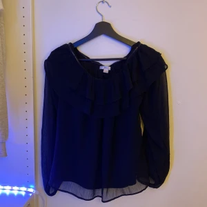 Mörkblå volangblus från Gina Tricot - Supersöt svart blus från Gina Tricot med volangdetaljer runt halsringningen. Blusen har långärmade, luftiga ärmar och är tillverkad i ett tunt, flowy material som ger en lätt och elegant känsla.