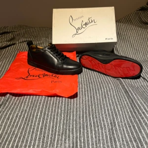Svarta sneakers från Christian Louboutin - Snygga svarta sneakers från Christian Louboutin i skinn med klassisk röd sula och diskret logga. Skorna har rund tå, svarta snören och stilren design som ger en lyxig vibe. Perfekta för dig som vill sticka ut med exklusiva detaljer. Sulorna är i nästan perfekt skick och resten av skon med skriv om andra detaljer eller funderingar.
