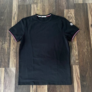 Svart Moncler t-shirt slim fit - Svart t-shirt från Moncler i slim fit med korta ärmar. T-shirten har Moncler-logga broderad på vänster ärm och snygga rödvita detaljer på ärmslut och halsringning. Tillverkad i mjuk bomull, perfekt för en clean och stilren look.