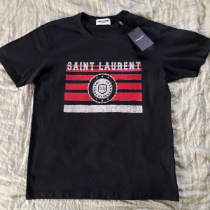 Svart Saint Laurent t-shirt med tryck - Svart t-shirt från Saint Laurent. Storlek M, köpt i frankrike! Ganska ”fitted” look. Helt ny kondition! Har haft i ca 3 månader men inte använts en enda gång.