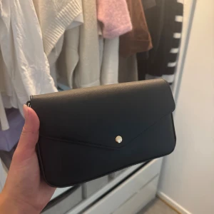 Svart kuvertväska från Don Donna - Stilren svart kuvertväska från Don Donna med guldfärgad kedja som axelrem. Väskan har ett elegant kuvertlock med knappstängning och är gjord i ett strukturerat syntetmaterial. Perfekt när du vill ha med det viktigaste och ändå se chic ut. Endast använd 1 gång helt som ny.