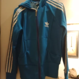Blå Adidas hoodie med vita ränder - Säljer en blå Adidas hoodie med klassiska vita ränder längs ärmarna och stor Adidas-logga på huvan. Tröjan har dragkedja framtill, huva och två fickor. Perfekt för dig som gillar sportig och streetwear-stil.