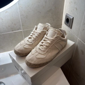 Maison Margiela sneakers i beige canvas - Maison Margiela sneakers i beige canvas med råa kanter och snygga detaljer. Skorna har snörning och en unik sula i flätad jute som ger en cool espadrillos-vibe. Perfekta för dig som gillar en avslappnad men trendig stil.