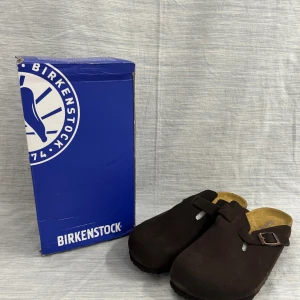 Bruna Birkenstock Boston i mocka - Säljer ett par mörkbruna Birkenstock Boston med ovandel i mjuk mocka och justerbart spänne i metall. Skorna har en klassisk rund tå och bekväm fotbädd i kork. Perfekta för dig som gillar stilren och chill look.