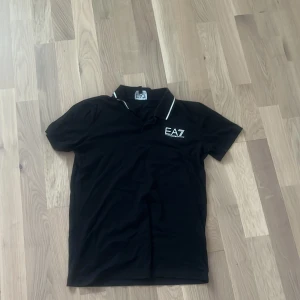 Svart EA7 t-shirt med krage - Svart t-shirt från EA7 Emporio Armani med vit logga på bröstet och vit kant på kragen. Klassisk polo-look med korta ärmar och mjukt material som känns skönt mot huden. Perfekt för dig som gillar stilrena och sportiga plagg.