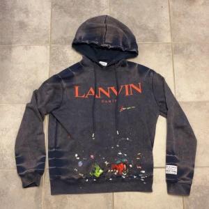 Lanvin x gallary dept hoodie - Säljer en mörkblå hoodie från Lanvin Paris med stor orange logga framtill och coolt färgstänksmönster nedtill. Tröjan har huva med snörning, lång ärm och en patch på vänster ärm. Perfekt för dig som gillar streetwear med konstnärlig vibe. Storlek M. Modellen på bilden är 185 cm och väger ca 70 kg