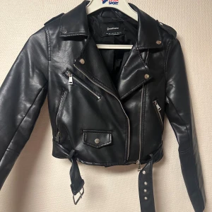 Svart skinnjacka från Stradivarius - Snygg svart skinnjacka från Stradivarius med klassisk bikerstil. Jackan har flera silvriga dragkedjor, och ett bälte i midjan. Croppad modell med långa ärmar och coola detaljer som ger en edgy vibe. 