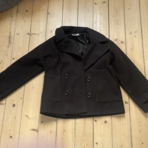 Svart skepparkavaj från Design by Si - Stilren svart skepparkavaj från Design by Si i kortare modell. Jackan har dubbelknäppning med stora knappar, bred krage och två rejäla fickor framtill. Perfekt för dig som gillar klassisk och tidlös stil.